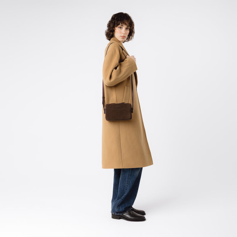 Model wearing STOW London's Camera bag in Chocolate Suede leather on her shoulder featuring gold-coated stainless steel hardware on a white background