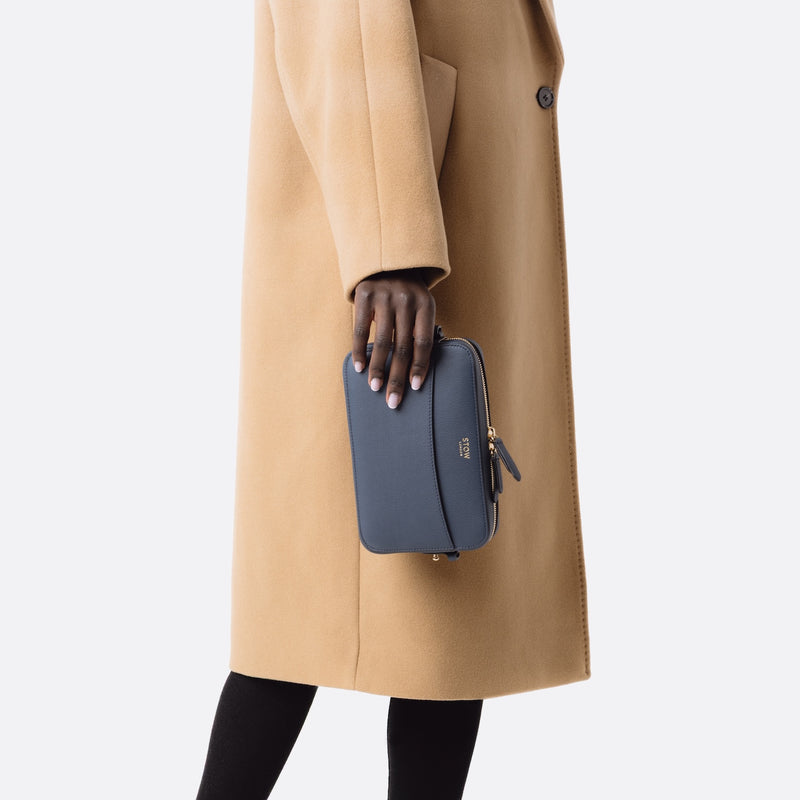Model holding STOW London's Camera bag in Navy Smooth leather in hand by her side featuring gold-coated stainless steel hardware on a white background
