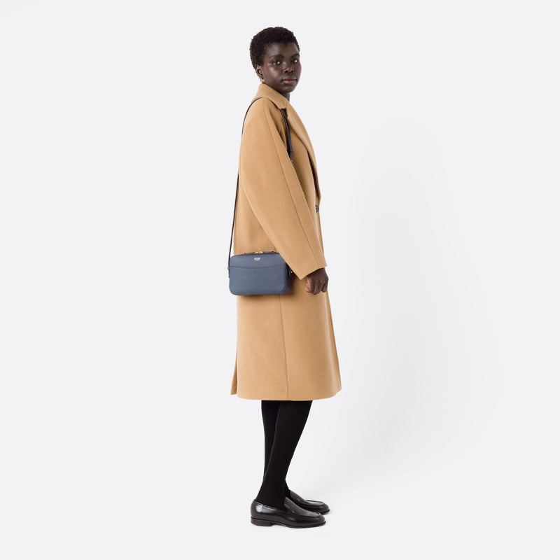 Model wearing STOW London's Camera bag in Navy Smooth leather on her shoulder with adjustable strap featuring gold-coated stainless steel hardware on a white background