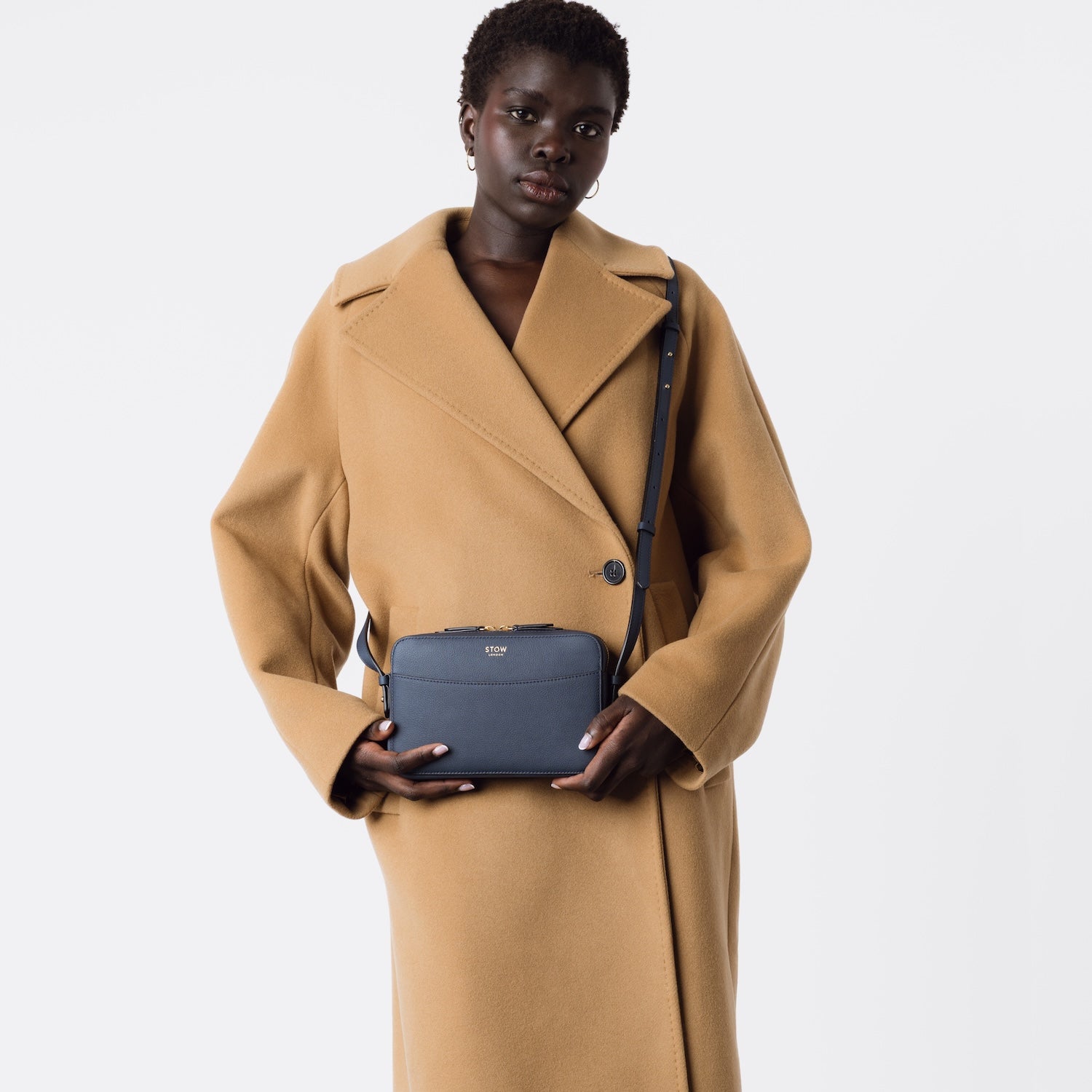 Model wearing STOW London's Camera bag in Navy Smooth leather crossbody with detachable strap featuring gold-coated stainless steel hardware on a white background