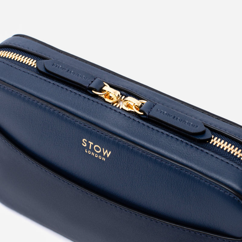 Close up of STOW London's Camera bag in Navy Smooth leather featuring gold-coated stainless steel hardware and brand engraved pull tabs on a white background	