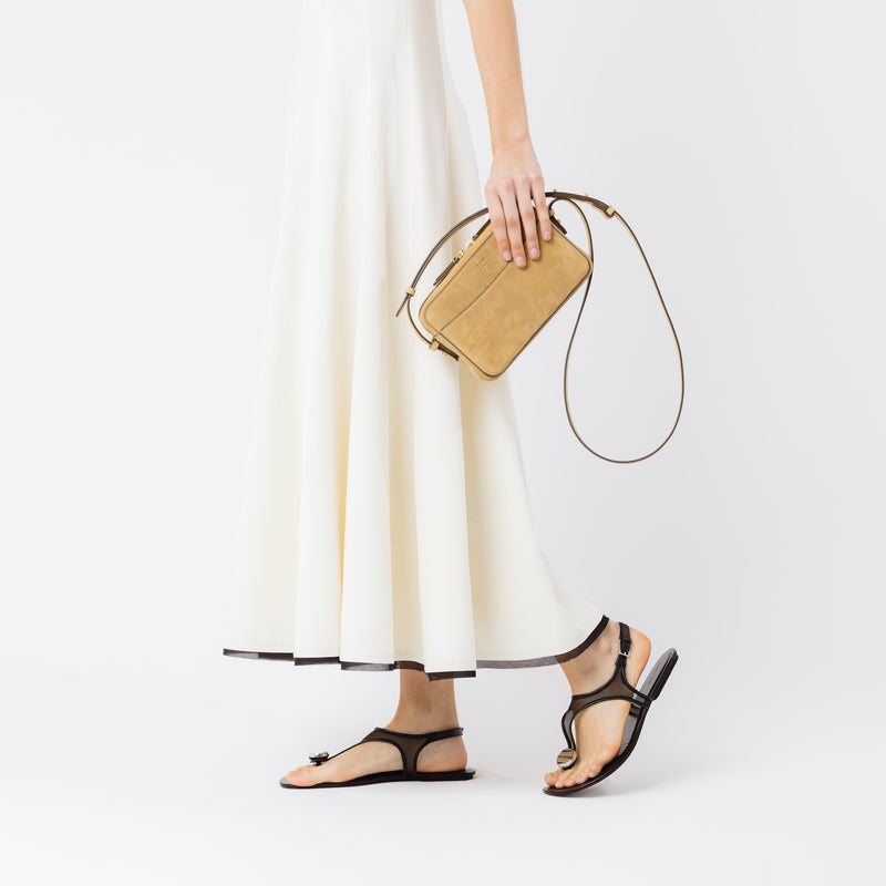 Model holding STOW London's Camera bag in Soft Sand Suede leather in her hand  at her side featuring gold-coated stainless steel hardware on a white background