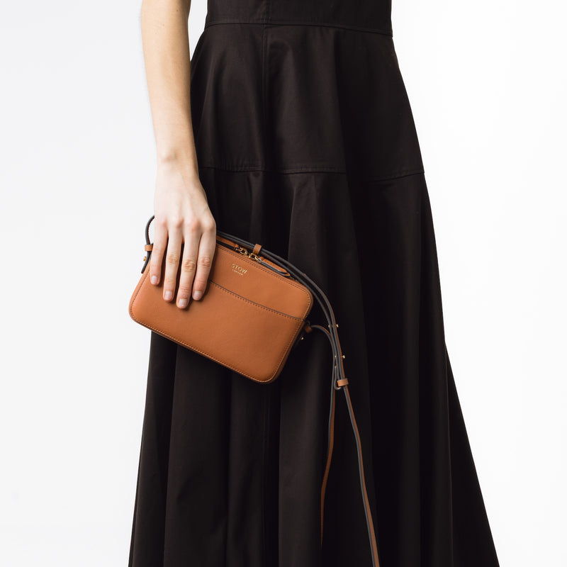 STOW London's Camera bag in Earth Tan smooth leather with detachable crossbody strap held by model at her side featuring gold-coated stainless steel hardware on a white background