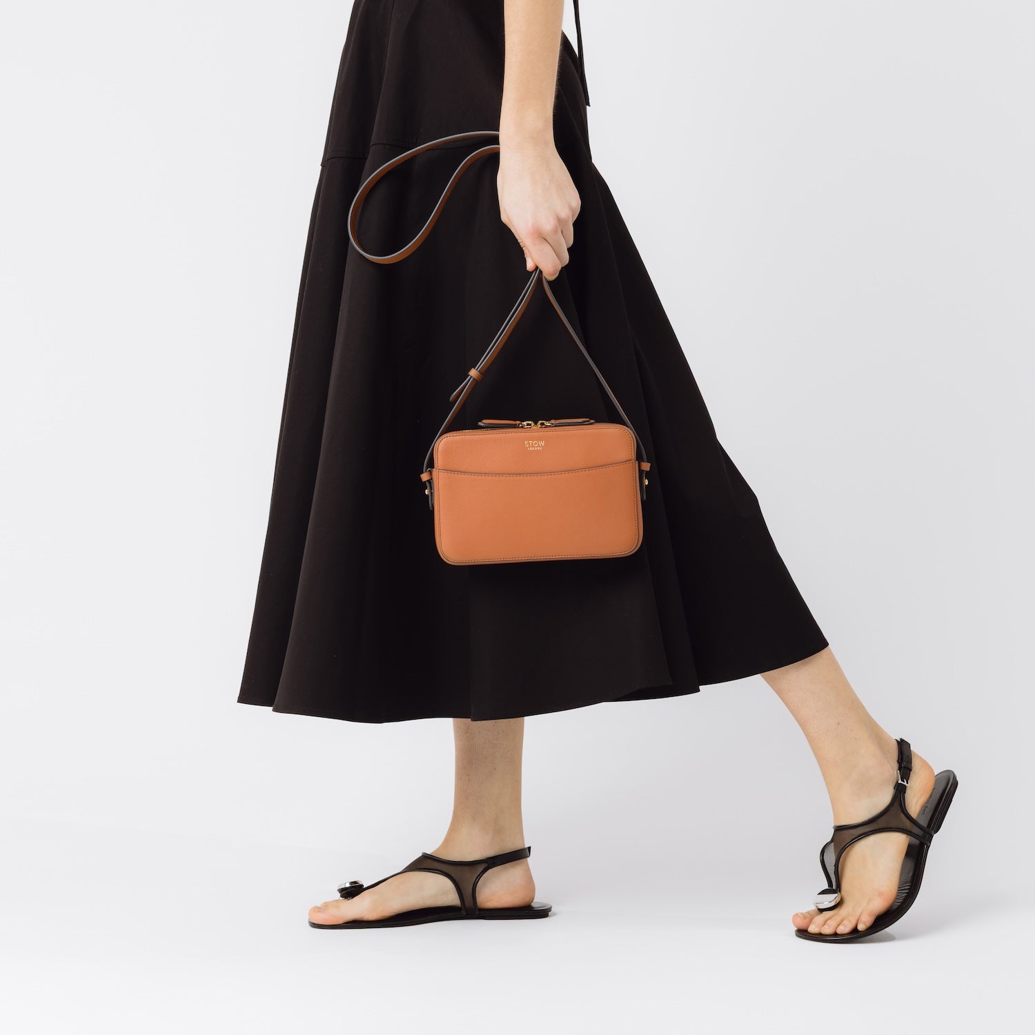 STOW London's Camera bag in Earth Tan smooth leather held by detachable crossbody strap in model's hand featuring gold-coated stainless steel hardware on a white background