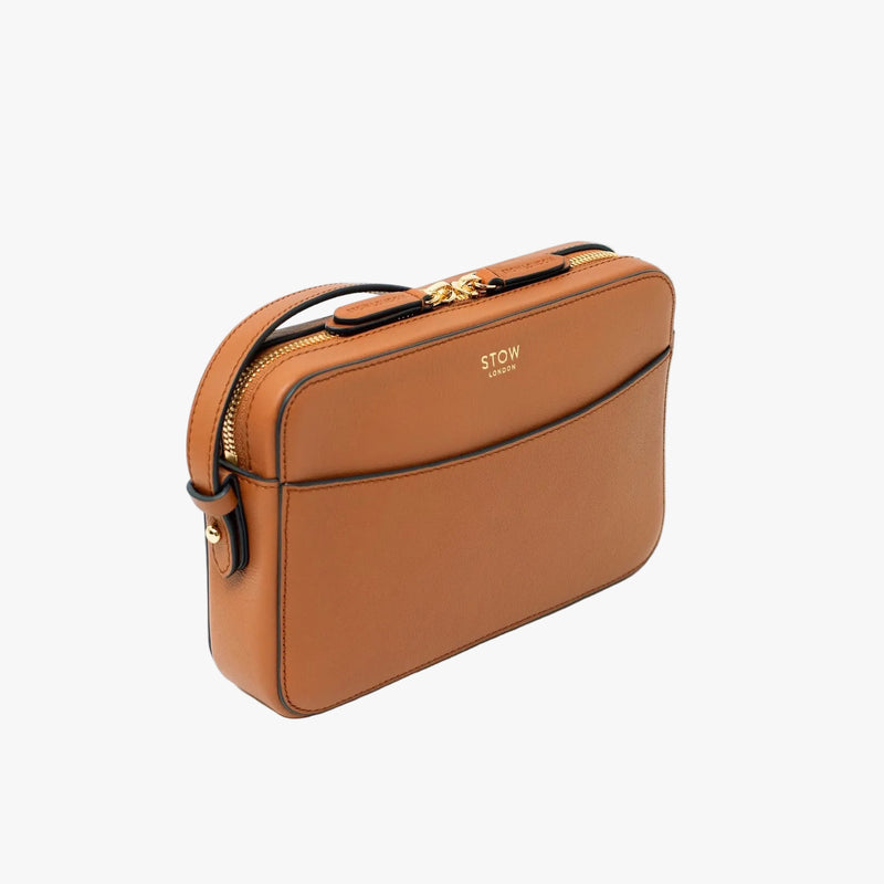Off-axis view of STOW London's Camera bag in Earth Tan pebbled leather featuring gold-coated stainless steel hardware on a light grey background 