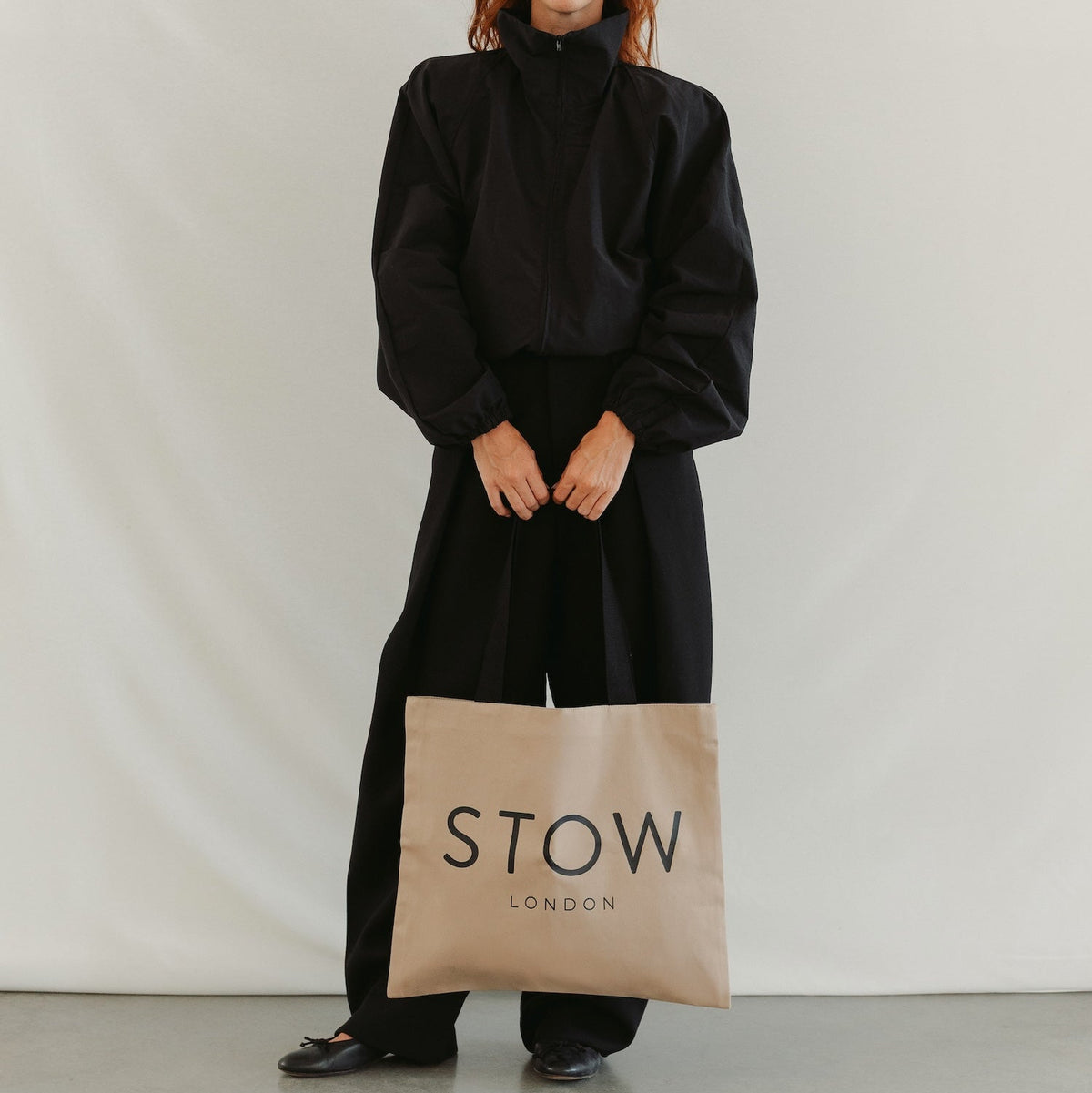 Model holding STOW London's Beige Canvas Tote bag featuring the brand name in black text, set against a plain light grey background.
