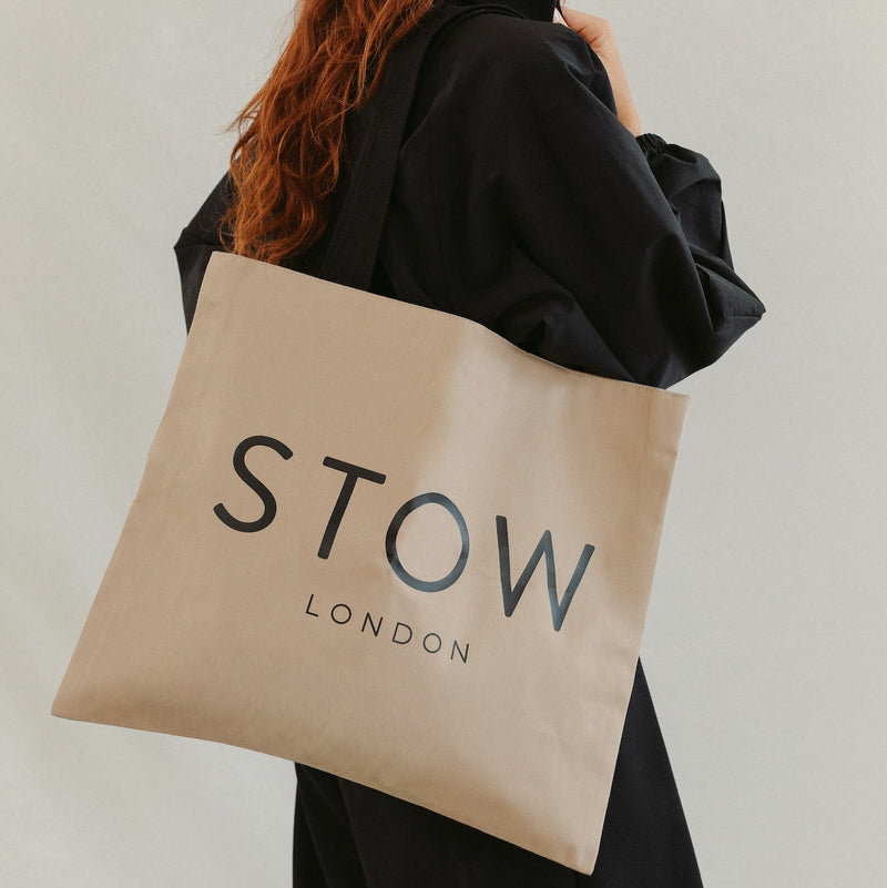 Model with long red hair carrying STOW London's Beige Canvas Tote bag featuring the brand name in black text, set against a plain light grey background.
