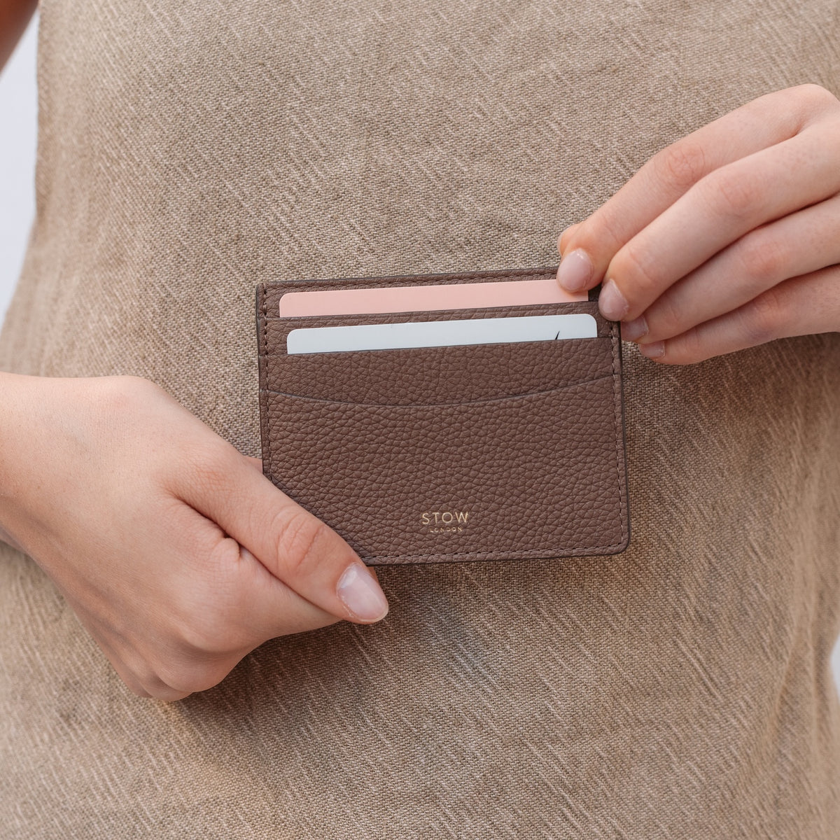 Model holding STOW London's Cardholder in Mocha pebbled leather featuring two cards and a small gold embossed logo on the front 