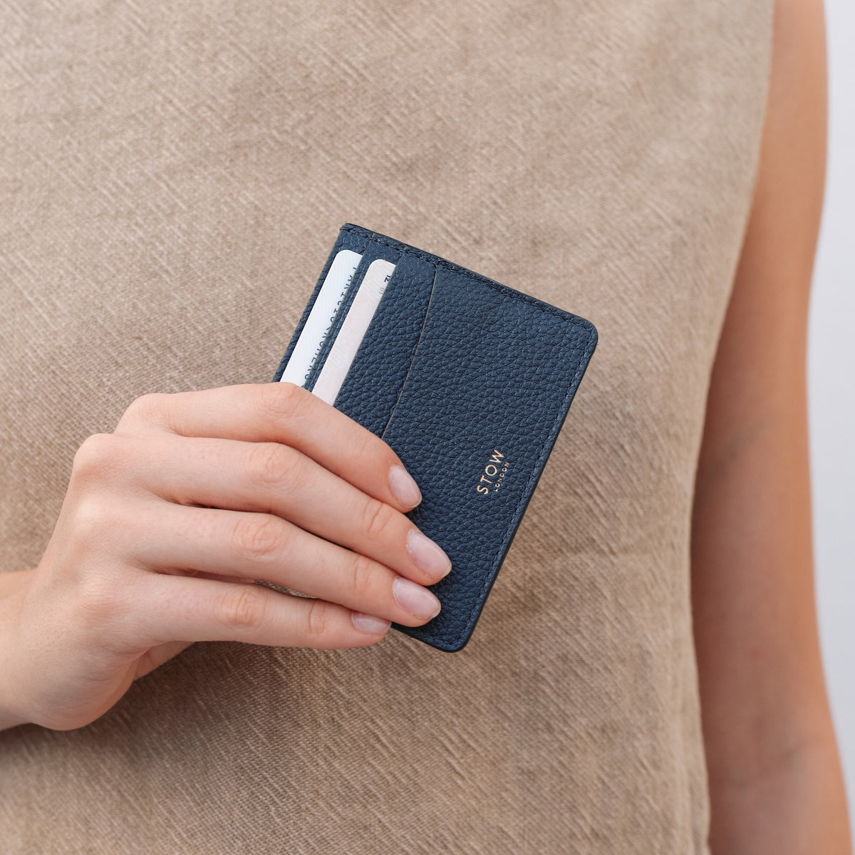 Model holding STOW London's Cardholder in Navy pebbled leather featuring two cards and a small gold embossed logo on the front 