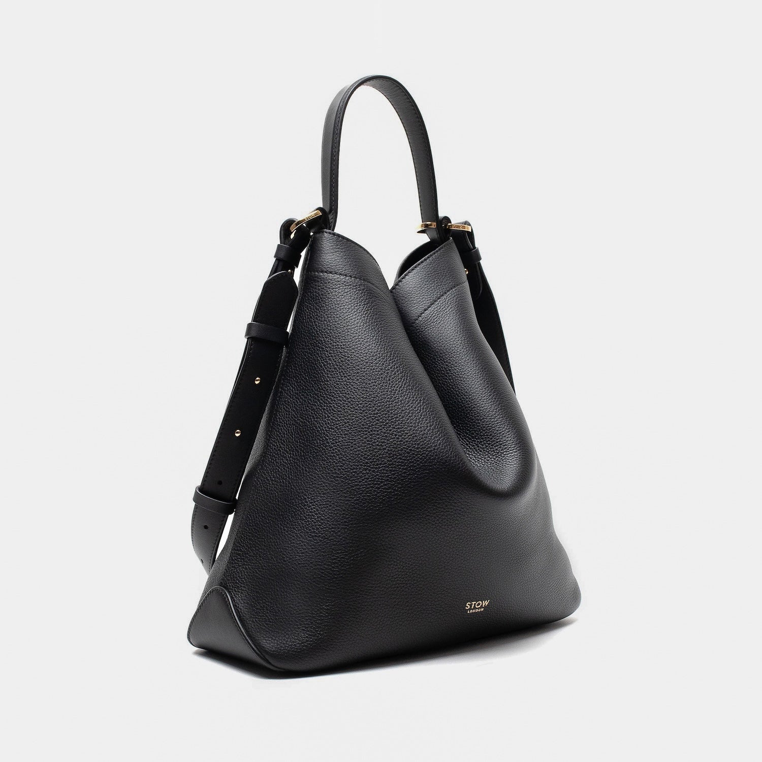 Off-axis shot of STOW London's Curve bag in Black pebbled leather in a triangular shape featuring gold-coated stainless steel hardware on a light grey background