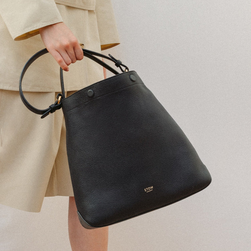 Model holding STOW London's Curve in Black leather featuring the detachable shoulder strap with gold hardware and details 