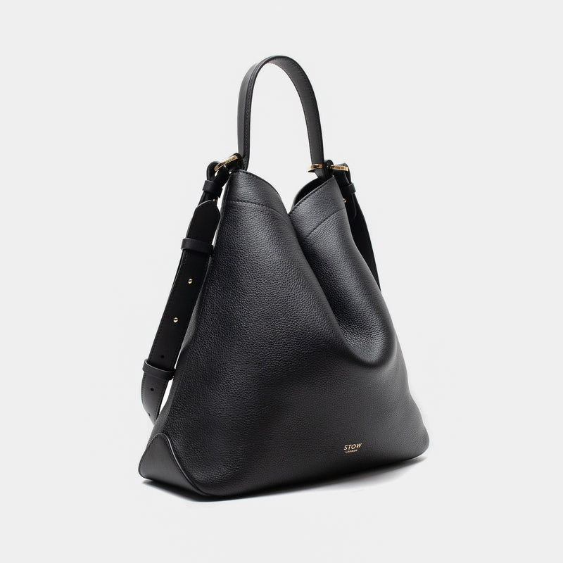 Off-axis shot of Stow London's Curve bag in Black pebbled leather in a triangular shape featuring gold-coated stainless steel hardware on a light grey background