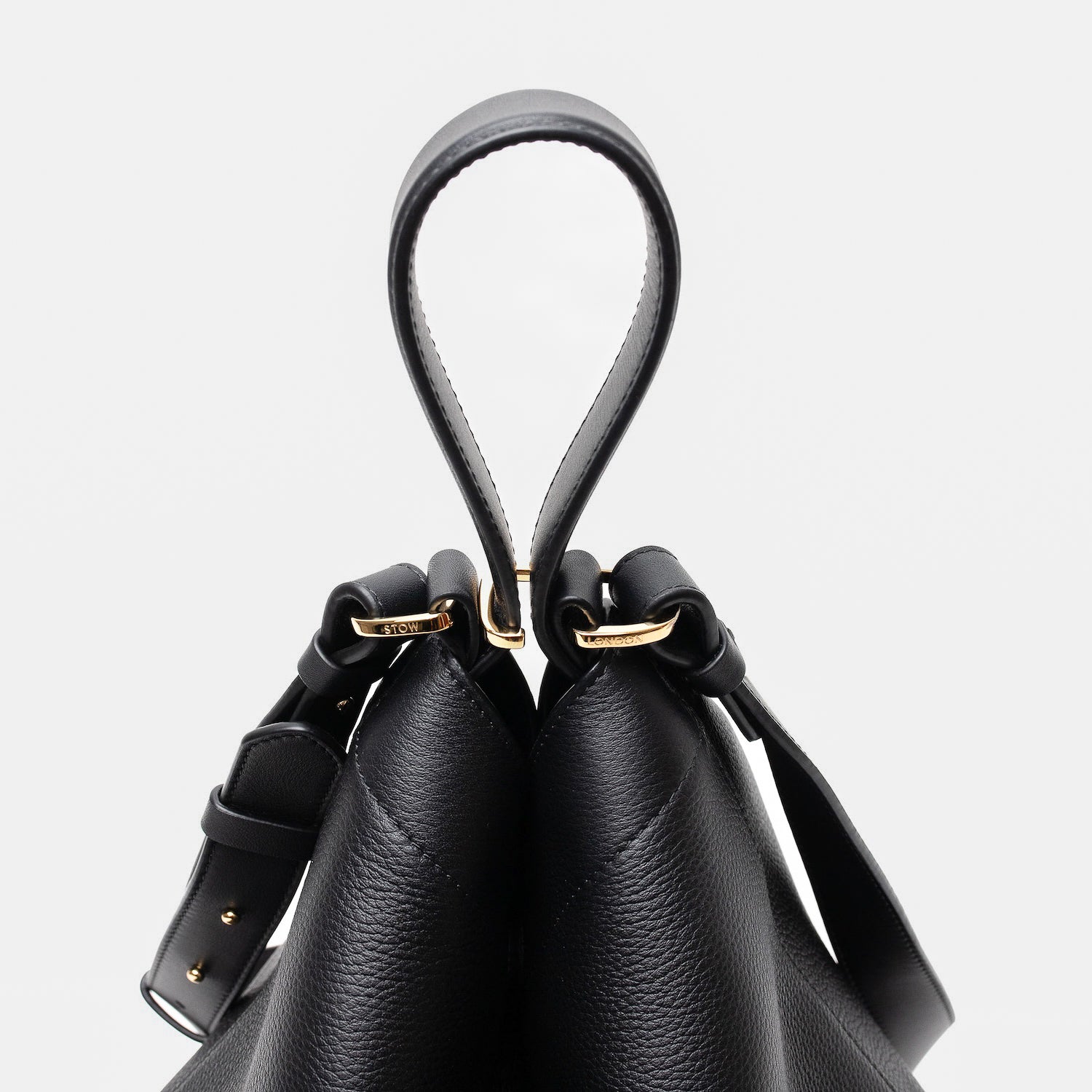 Close up of the handles on Stow London's Curve bag in Black pebbled leather featuring gold-coated stainless steel hardware and clip and detachable strap on a light grey background