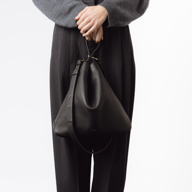 Model holding Stow London's Curve bag in Black pebbled leather by the handle at her front featuring gold-coated stainless steel hardware on a light grey background