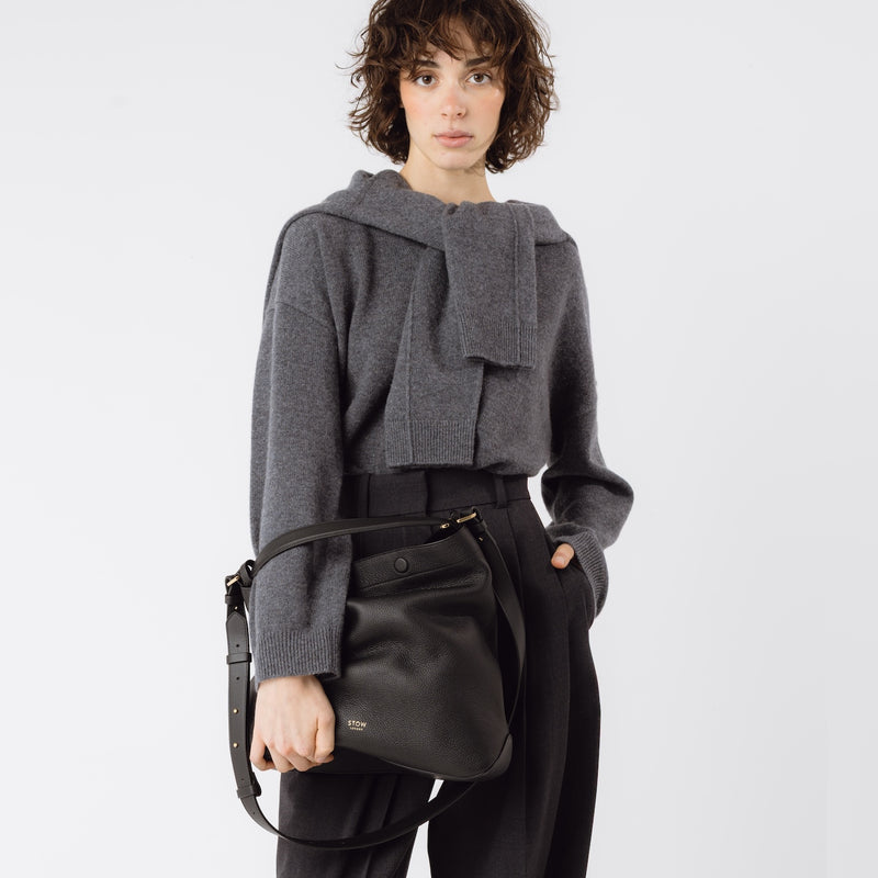 Model holding Stow London's Curve bag in Black pebbled leather featuring gold-coated stainless steel hardware on a light grey background