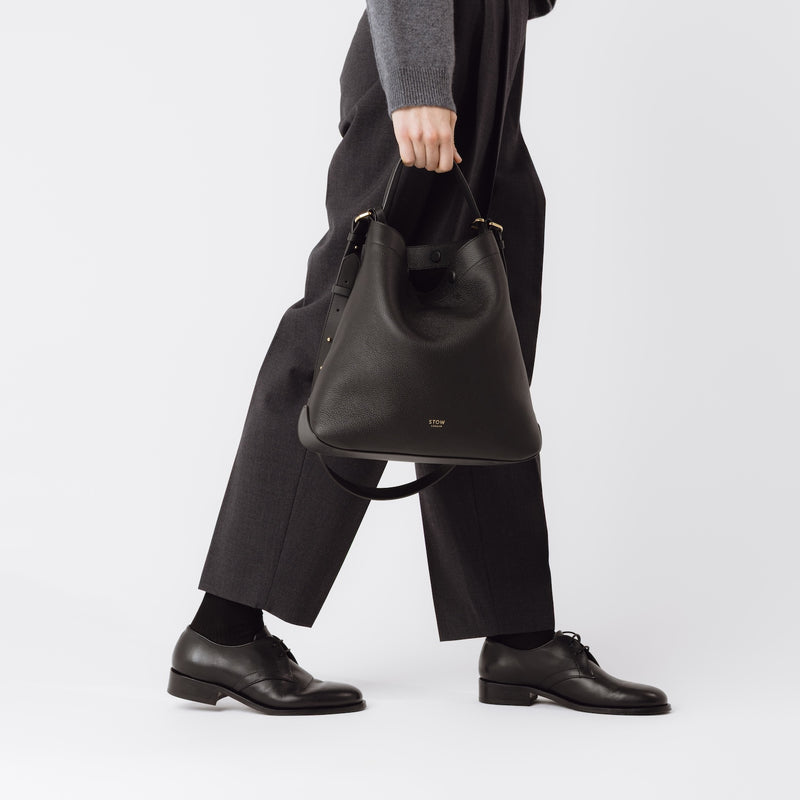 Model holding Stow London's Curve bag in Black pebbled leather at her side featuring gold-coated stainless steel hardware on a light grey background