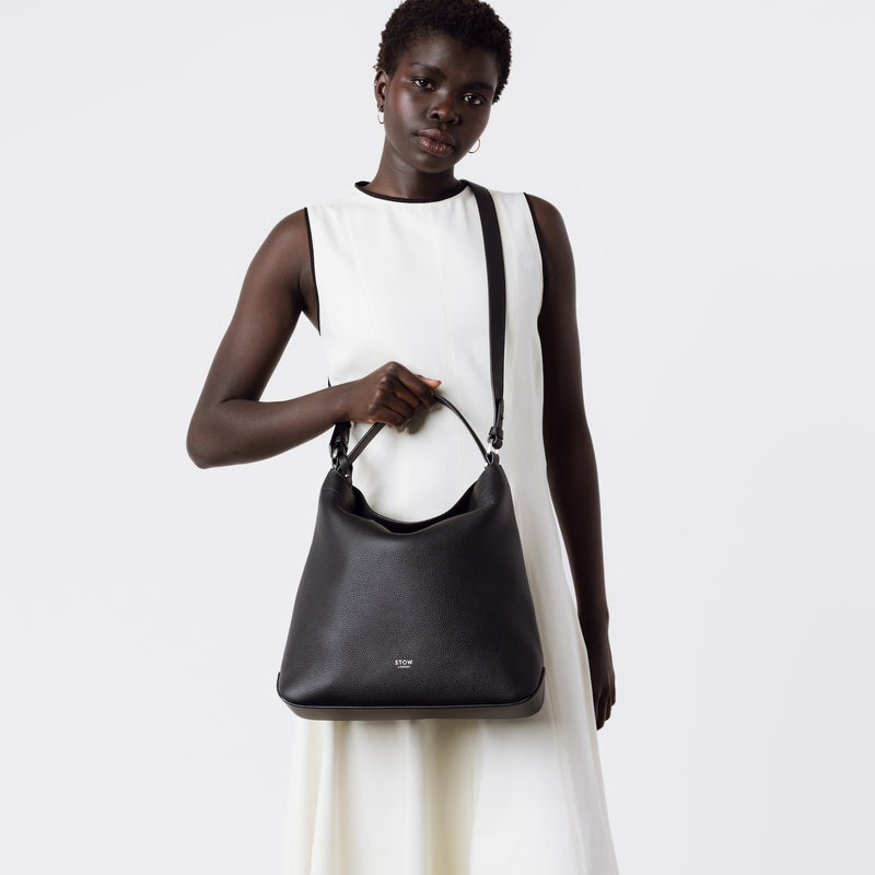 Model wearing Stow London's Curve bag in Black pebbled leather crossbody featuring silver stainless steel hardware on a light grey background