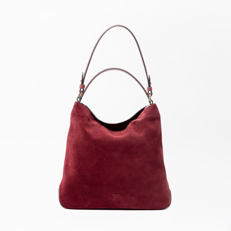 STOW London's Curve in Burgundy suede leather on a light grey background featuring the detachable shoulder strap and top handle with gold hardware