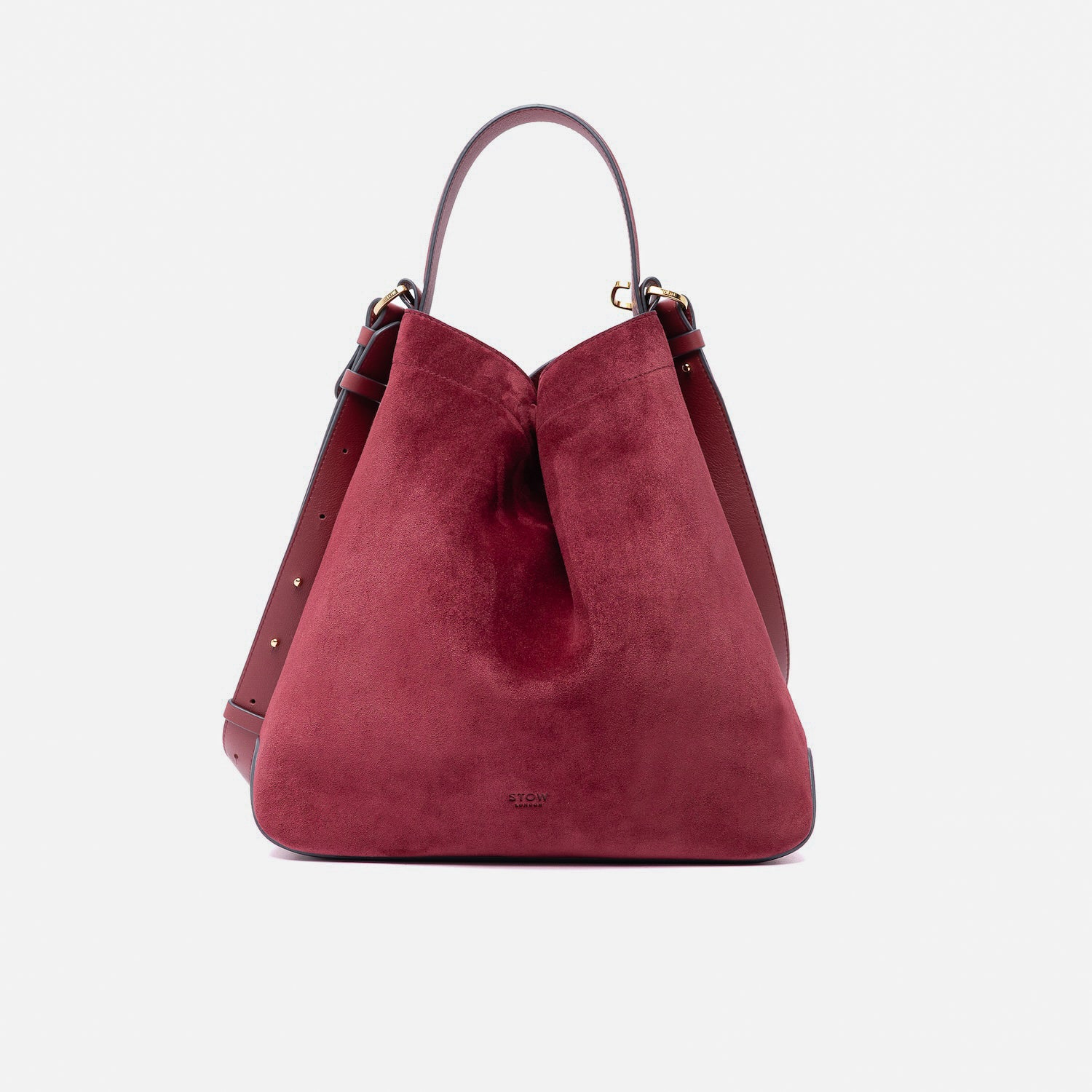 Stow London's Curve bag in Burgundy suede leather shaped in a triangular form featuring gold-coated stainless steel hardware on a light grey background