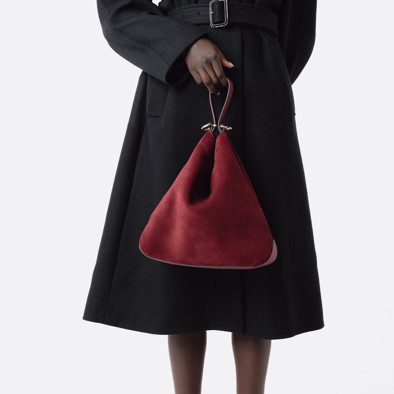 Model holding Stow London's Curve bag in Burgundy suede leather by handle at her front featuring gold-coated stainless steel hardware on a light grey background