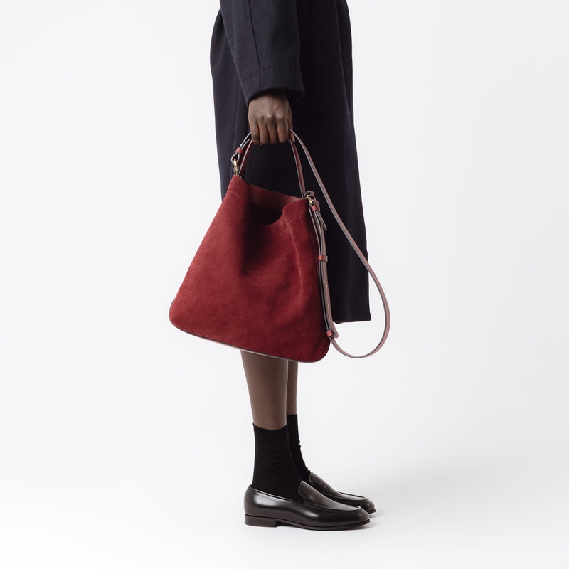 Model holding Stow London's Curve bag in Burgundy suede leather at her side featuring gold-coated stainless steel hardware on a light grey background