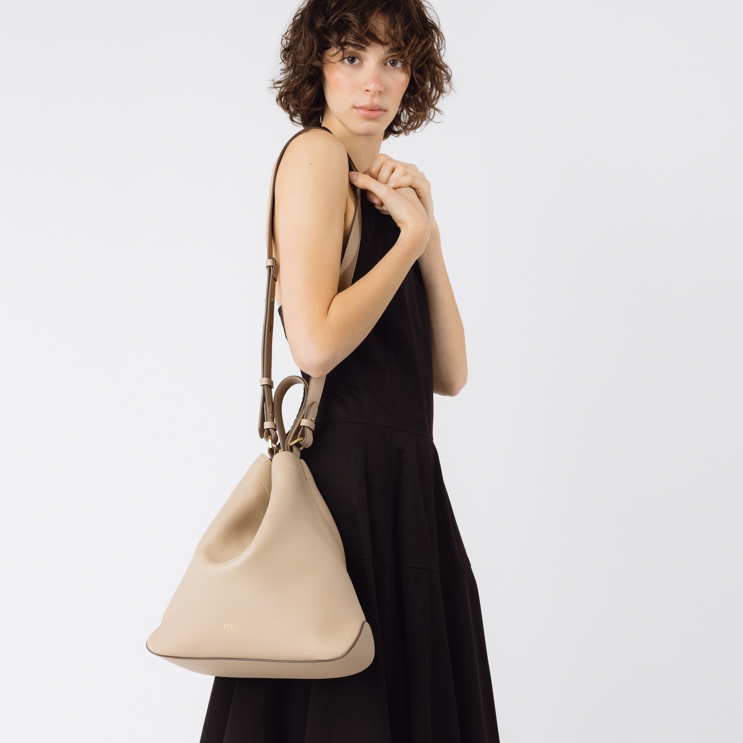 STOW London's Curve bag in Camel leather worn over shoulder with detachable strap featuring gold-coated stainless steel hardware on a white background