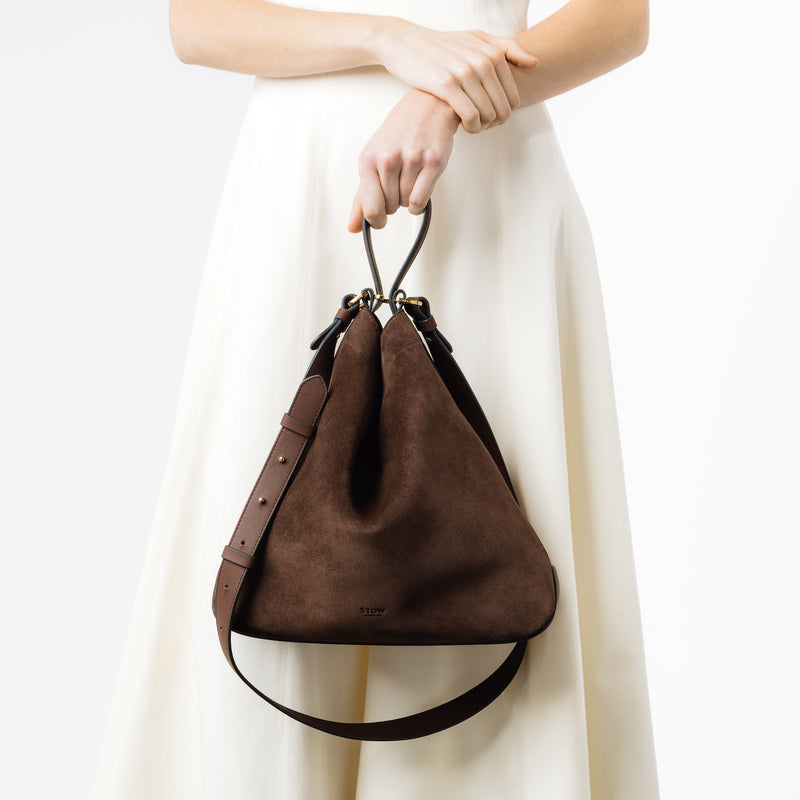 Model holding STOW London's Curve bag in Chocolate suede leather at her front featuring gold-coated stainless steel hardware on a light grey background
