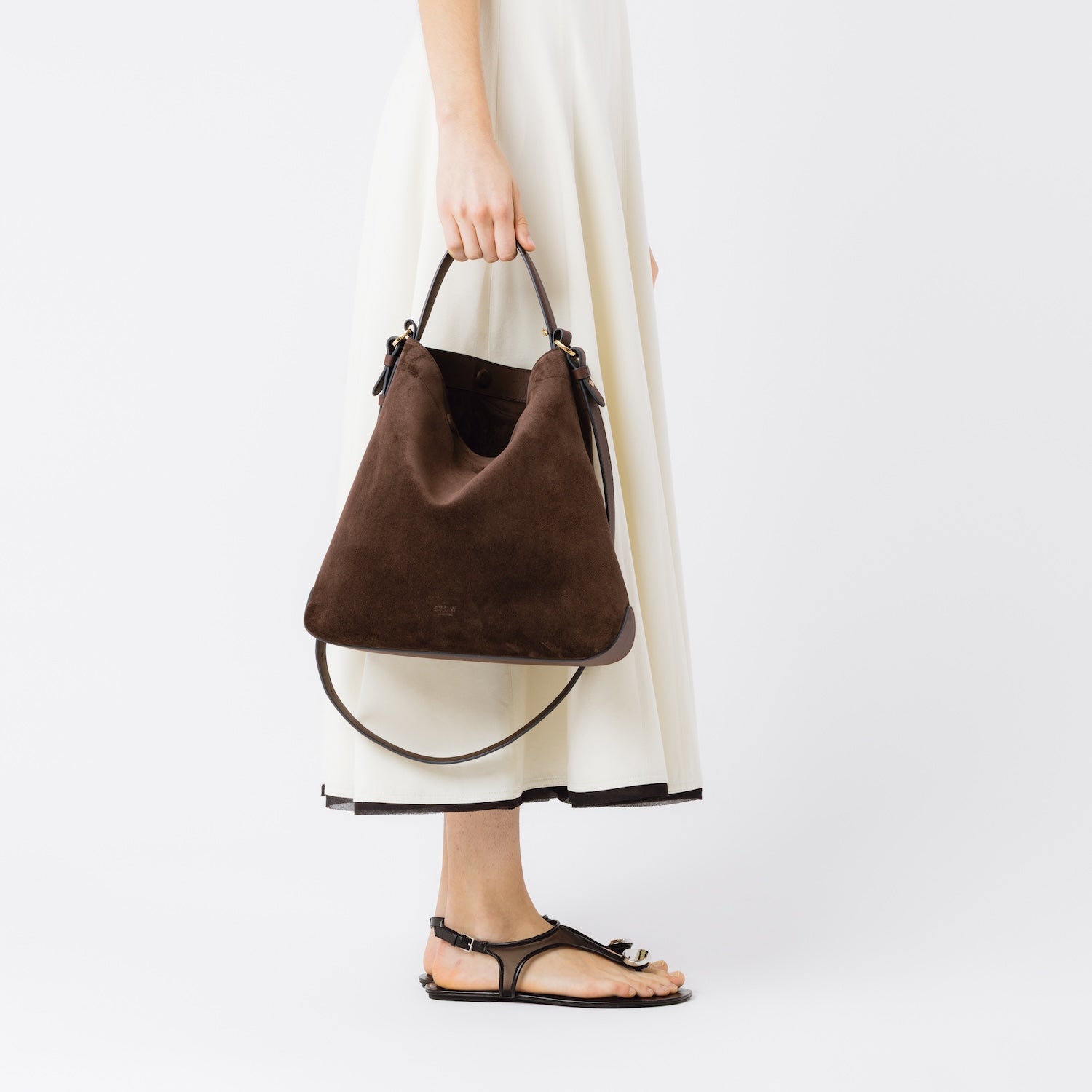 Model holding STOW London's Curve bag in Chocolate suede leather at her side featuring gold-coated stainless steel hardware on a light grey background