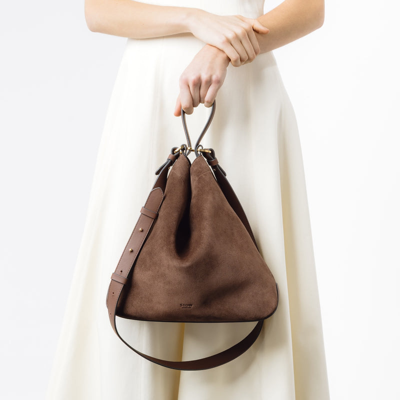 Model holding Stow London's Curve bag in Chocolate suede leather at her front featuring gold-coated stainless steel hardware on a light grey background