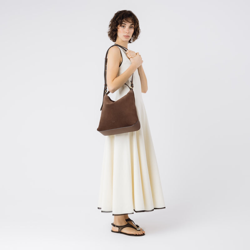 Model wearing Stow London's Curve bag in Chocolate suede leather on her shoulder featuring gold-coated stainless steel hardware on a light grey background