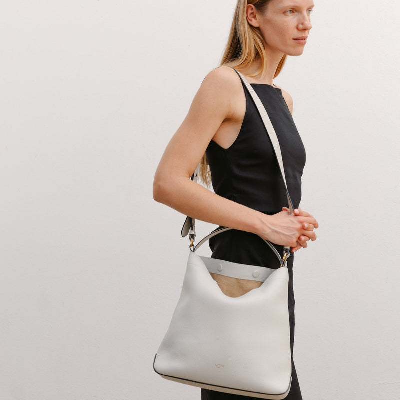 Model wearing STOW London's Curve bag in Cloud Grey pebbled leather by its crossbody strap featuring gold-coated stainless steel hardware 