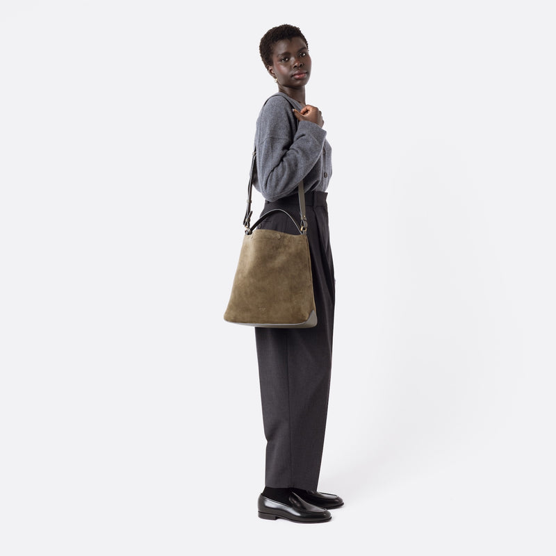 Model wearing Stow London's Curve bag in Dark Green suede leather on her shoulder featuring gold-coated stainless steel hardware and the detachable crossbody strap on a white background