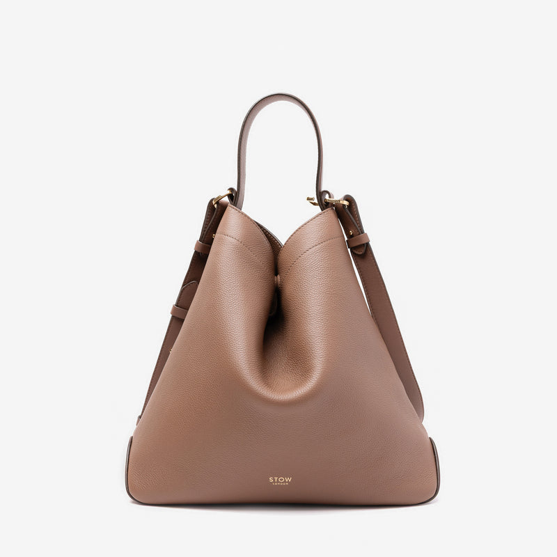 Stow London's Curve bag in Mocha pebbled leather shaped in a triangular form featuring gold-coated stainless steel hardware on a light grey background