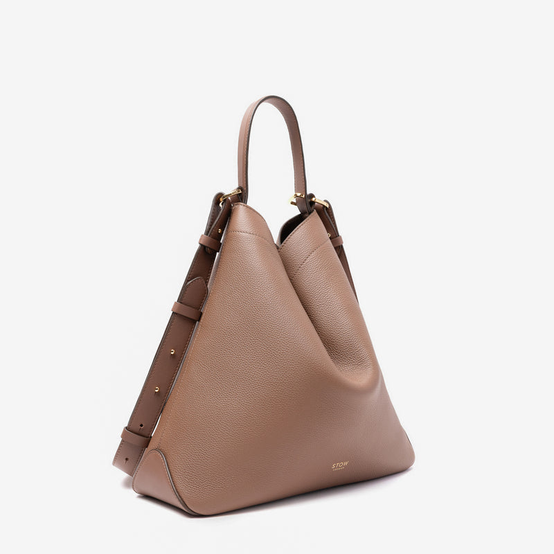 Off-axis view of Stow London's Curve bag in Mocha pebbled leather in a triangular form featuring gold-coated stainless steel hardware on a light grey background