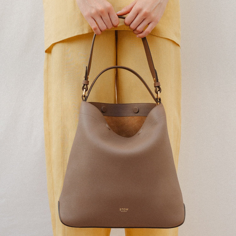 Model holding STOW London's Curve in Mocha leather featuring the detachable shoulder strap and top handle with gold hardware and details 