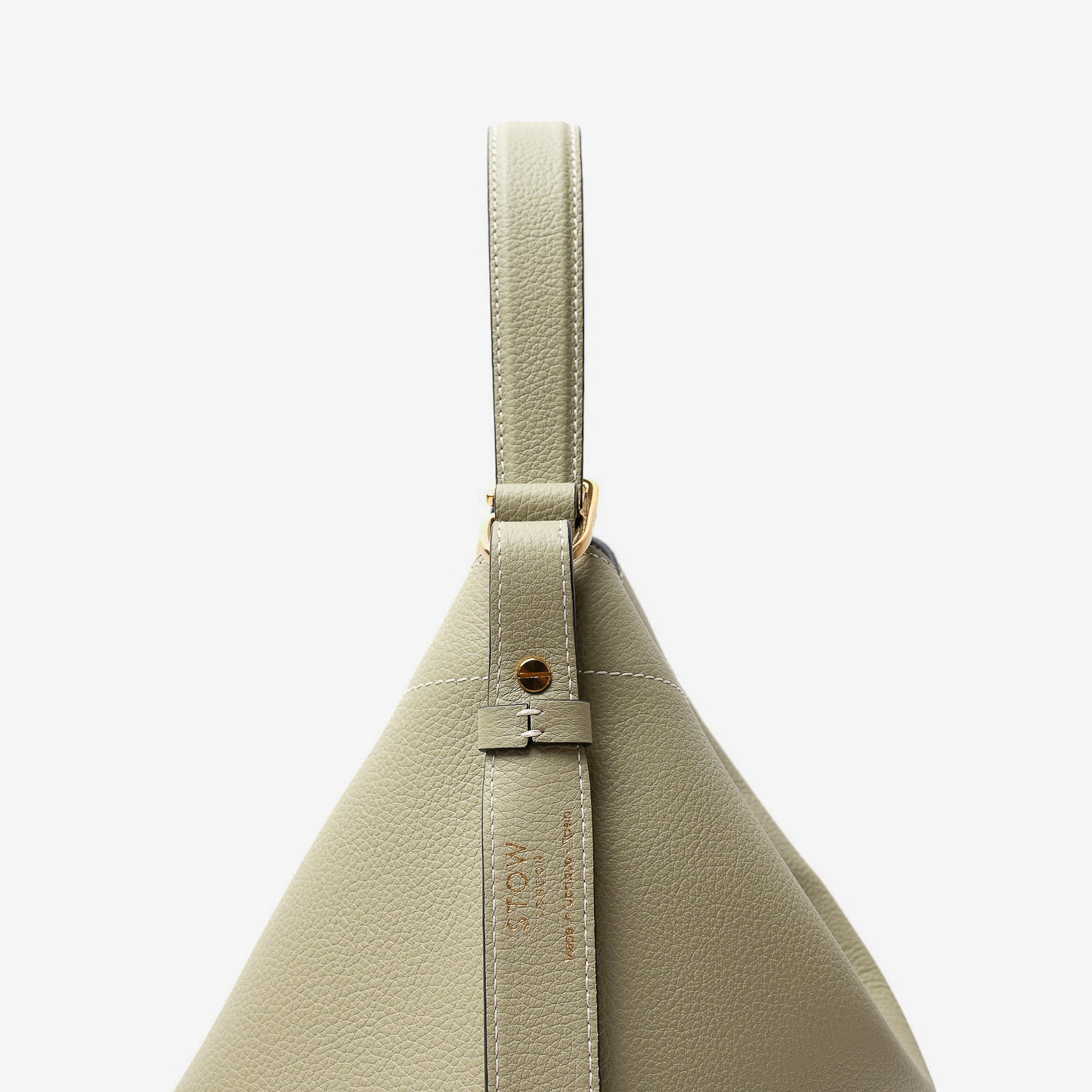 Close up of the handle and strap attachment on STOW London's Curve bag in Pale Green pebbled leather featuring gold-coated stainless steel hardware and clip and detachable strap on a light grey background