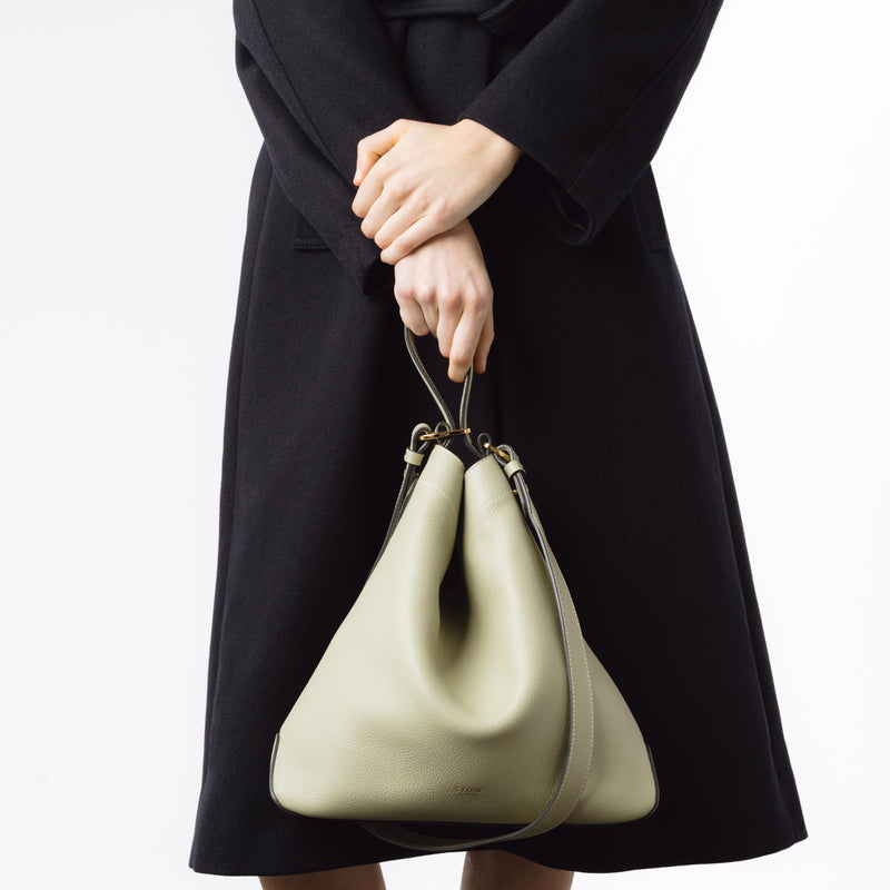 Model holding Stow London's Curve bag in Pale Green pebbled leather by the top handle featuring gold-coated stainless steel hardware on a light grey background