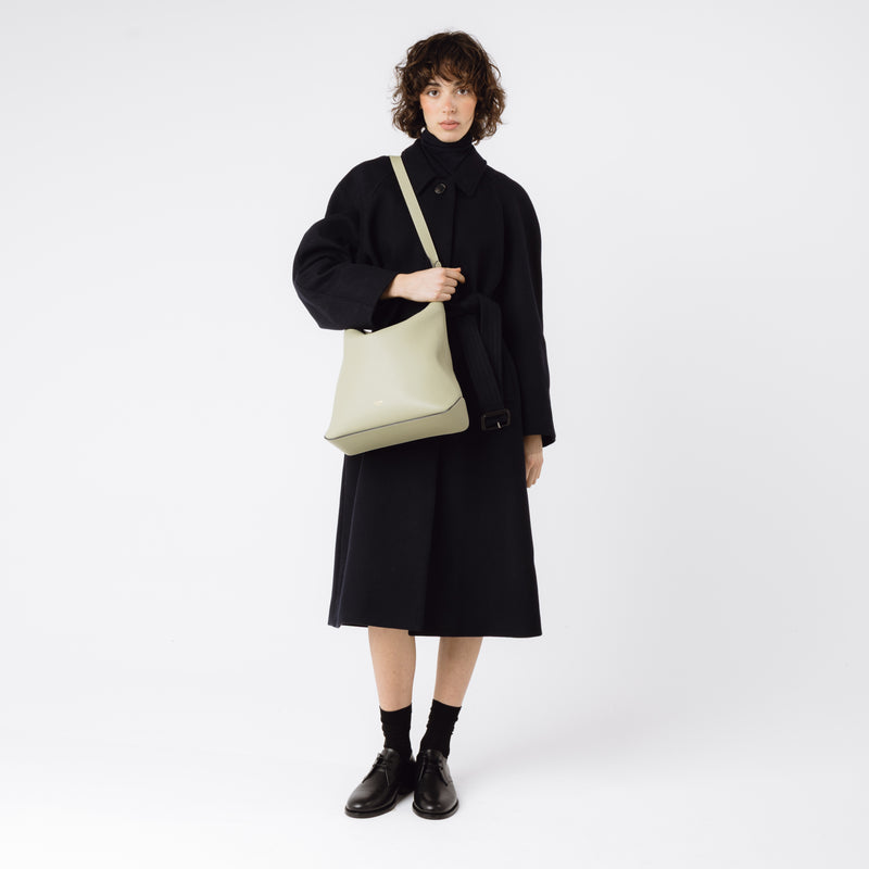 Model wearing Stow London's Curve bag in Pale Green pebbled leather on her shoulder featuring gold-coated stainless steel hardware on a light grey background