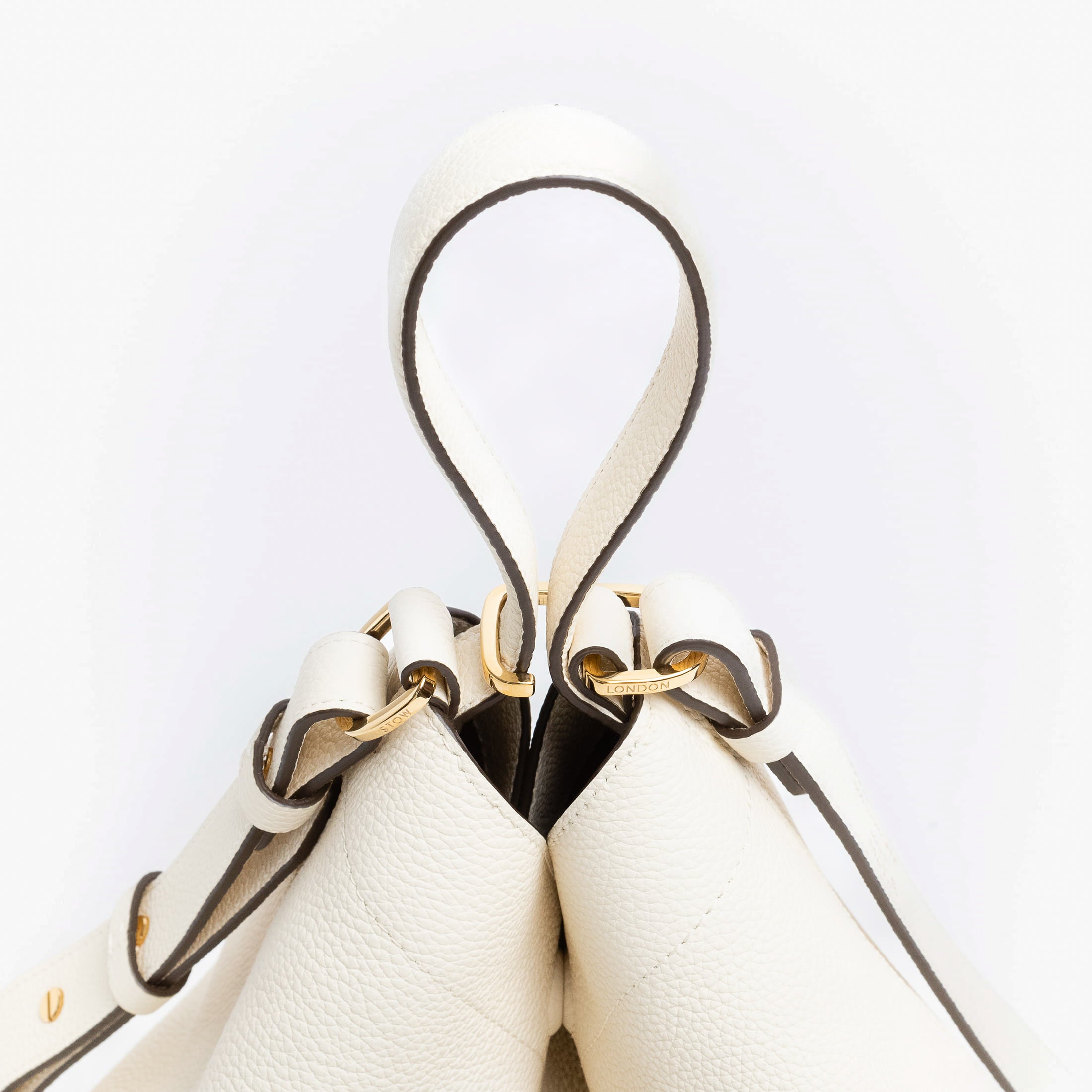 Close up of the handles on Stow London's Curve bag in Spring Moon pebbled leather featuring gold-coated stainless steel hardware and clip and detachable strap on a light grey background