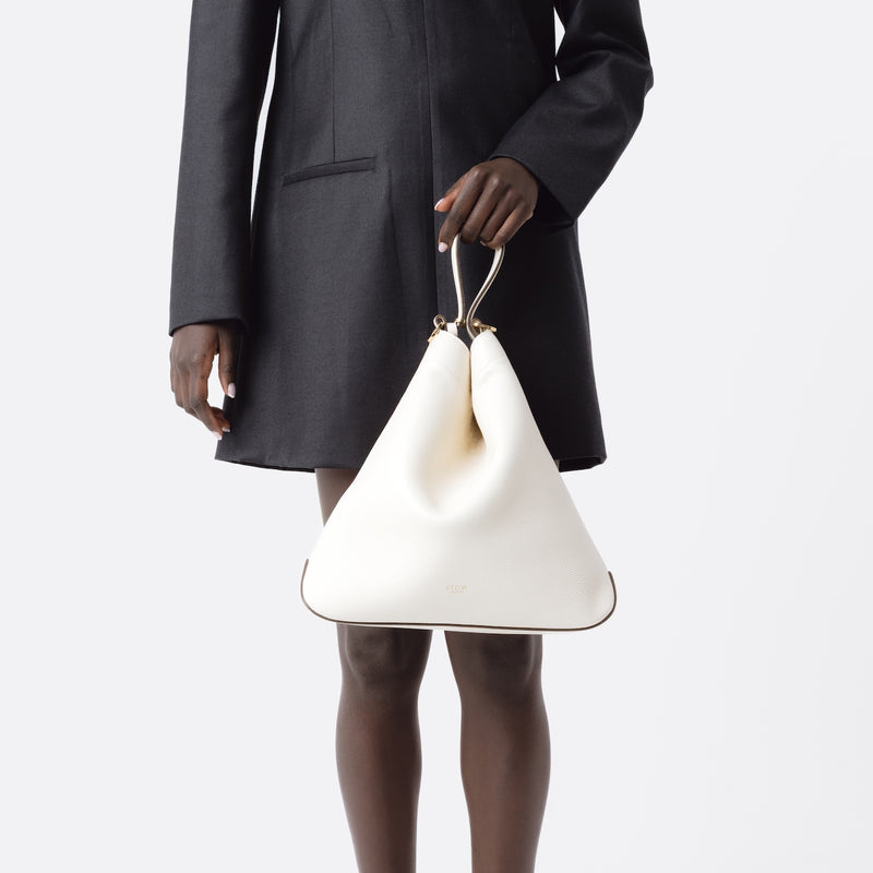 Model holding Stow London's Curve bag in Spring Moon pebbled leather at her front featuring gold-coated stainless steel hardware on a light grey background