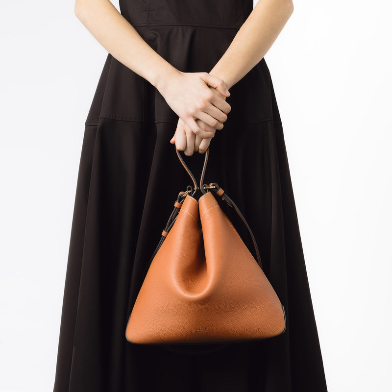 Model holding STOW London's Curve bag in Earth Tan by its top handle at her front featuring gold-coated stainless steel hardware on a white background 