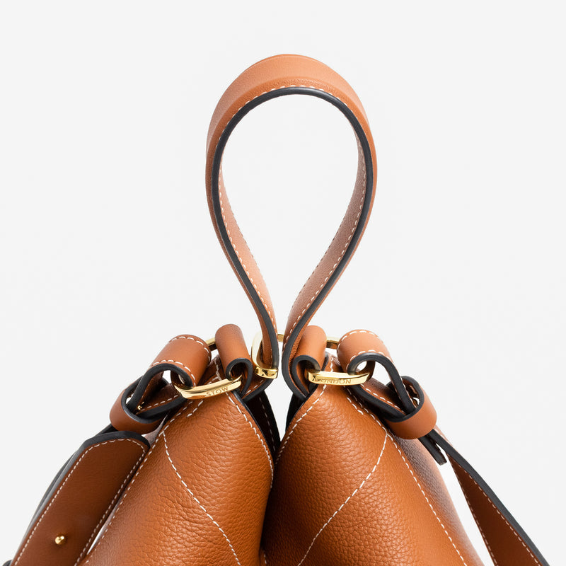 Close up of the handles on Stow London's Curve bag in Earth Tan pebbled leather featuring gold-coated stainless steel hardware and clip and detachable strap on a light grey background