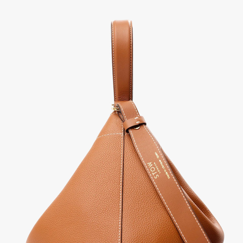 Side view of STOW London's Curve Bag in Earth tan pebbled leather and detachable crossbody strap on a light grey background featuring light gold embossed logo