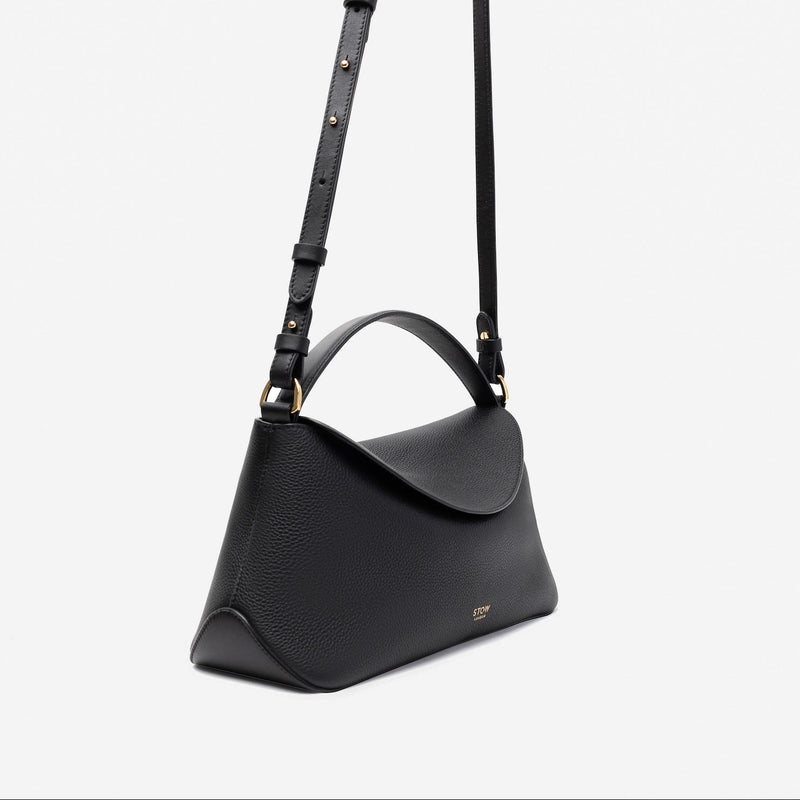 Off-axis view of STOW London's East West in Black pebbled leather and detachable crossbody strap featuring gold-coated stainless steel hardware on a light grey background
