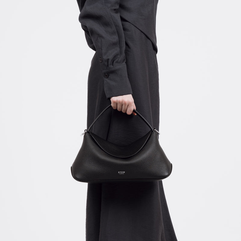 Close shot of model holding STOW London's East West bag in Black pebbled and smooth leather by its top handle at her side featuring silver stainless steel hardware on a white background