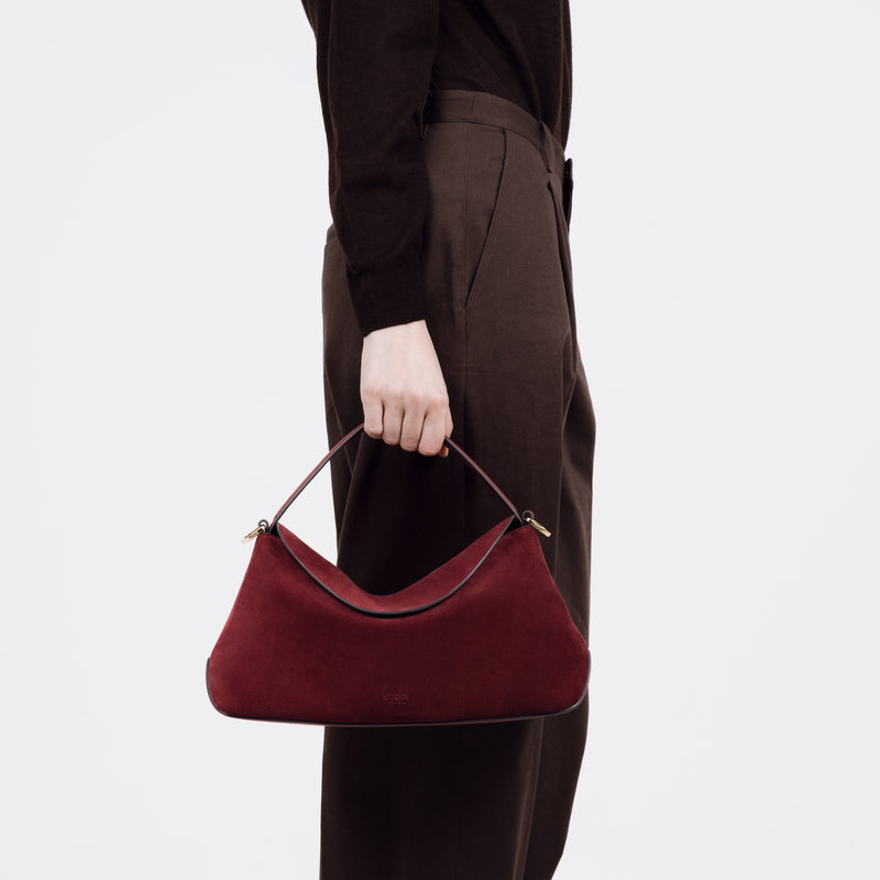 Model holding STOW London's East West bag in Burgundy Suede leather by the top handle at her side featuring gold-coated stainless steel hardware on a white background