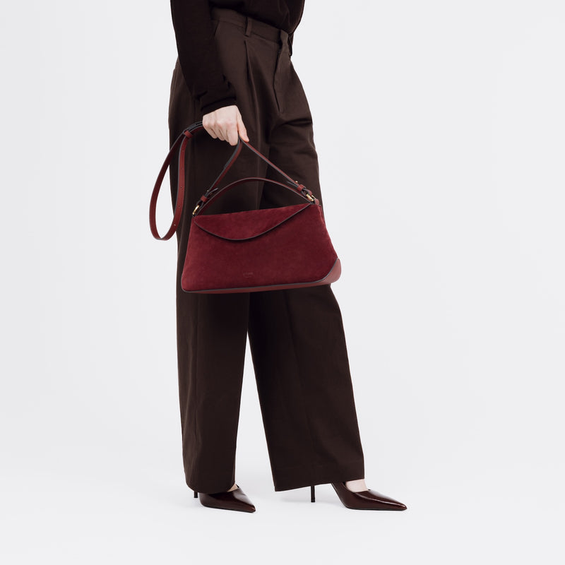 Model holding STOW London's East West bag in Burgundy Suede leather by its detachable crossbody strap at her side featuring gold-coated stainless steel hardware on a white background
