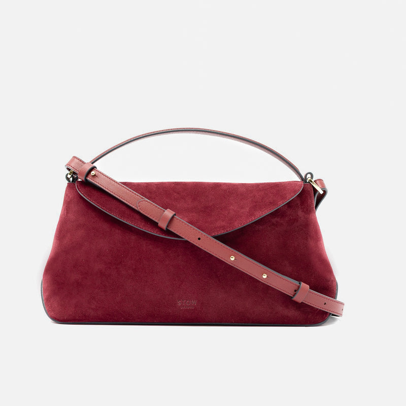 STOW London's East West in Burgundy suede leather and detachable crossbody strap featuring gold-coated stainless steel hardware on a light grey background