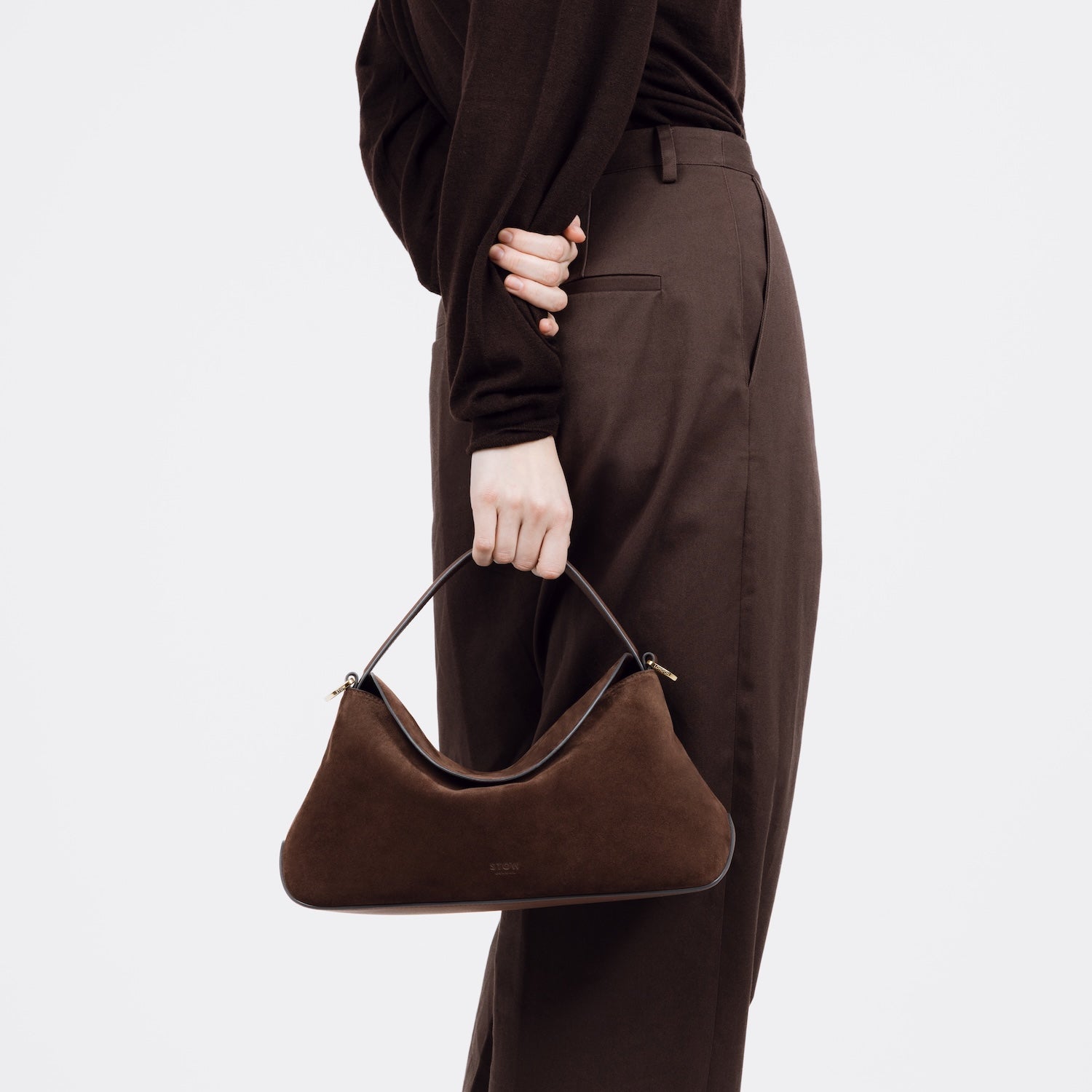 Model holding STOW London's East West bag in Chocolate Suede leather by the top handle behind her featuring gold-coated stainless steel hardware on a white background