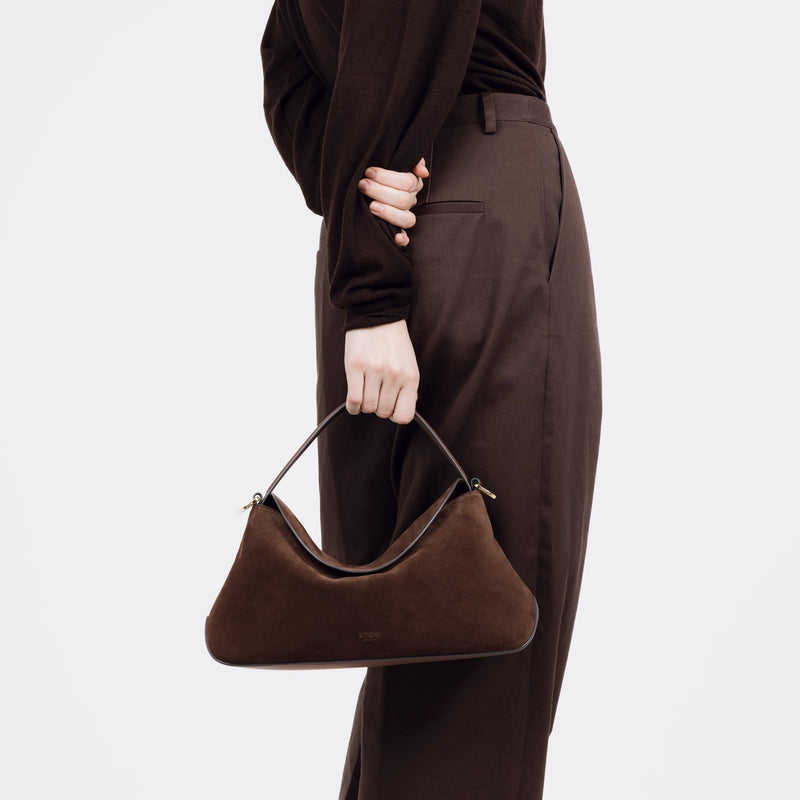 Model holding STOW London's East West bag in Chocolate Suede leather by the top handle featuring gold-coated stainless steel hardware on a white background